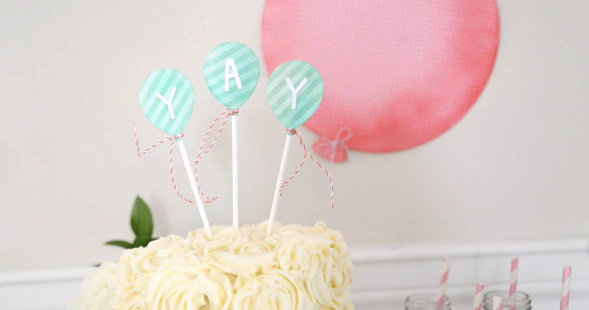 Birthday cake with balloon cards sticking out of it that say "yay"